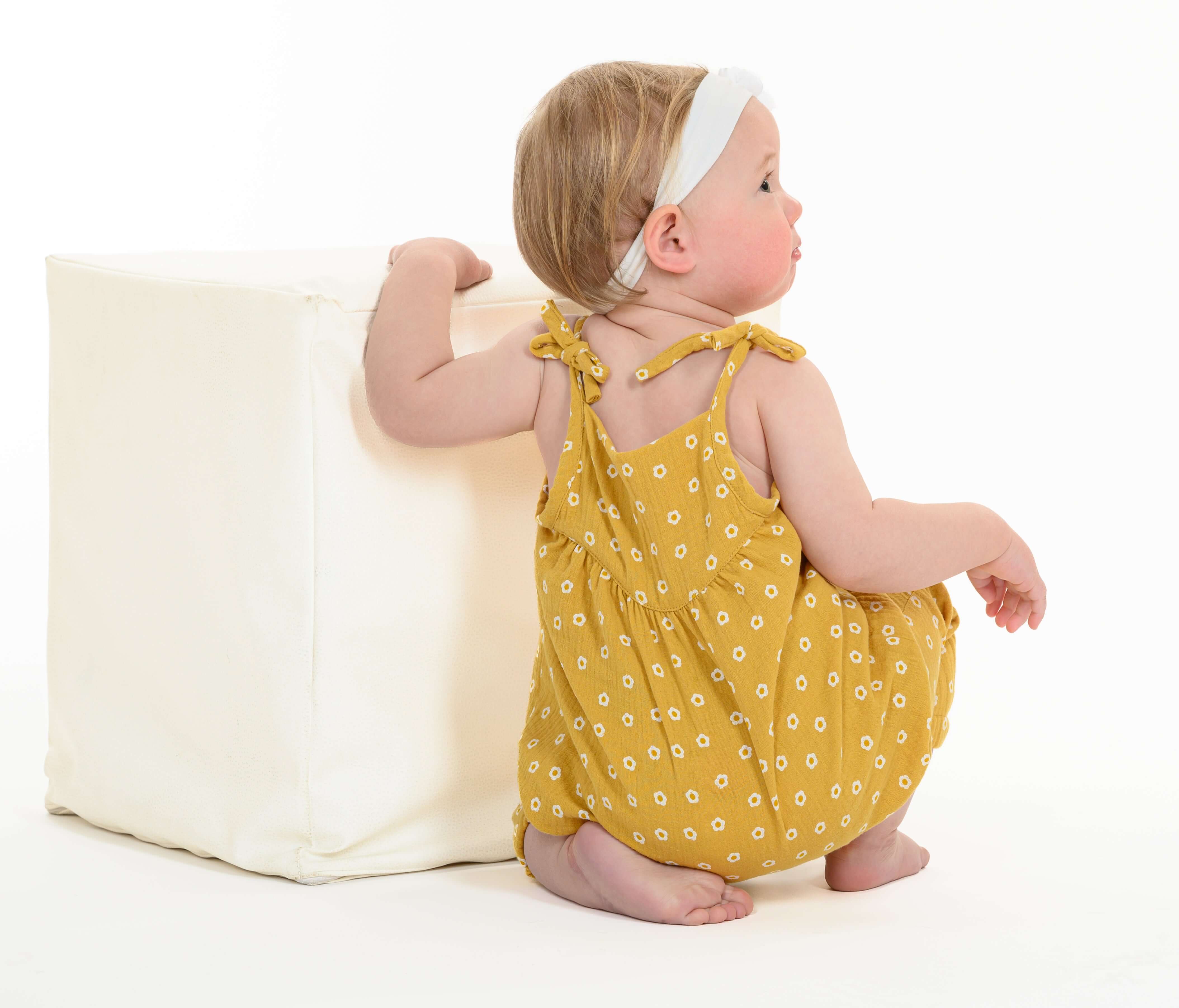 Schnittmuster Baby Overall Jumpsuit – Kleinkind im gelben Overall mit Blumenmuster, sitzt auf einem weißen Würfel.